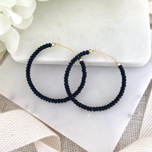 May include: A pair of black beaded hoop earrings with gold-colored accents. The earrings are displayed on a white marble surface with a white flower and a cream-colored ribbon in the background. The hoops are approximately 2 inches in diameter.
