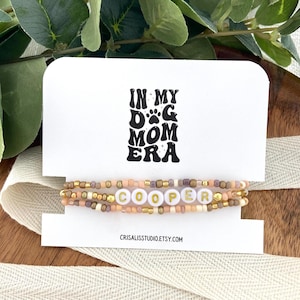 May include: A beaded bracelet featuring small tan, gold, and mauve beads with white letter beads spelling COOPER. The bracelet is displayed on a white card printed with the text IN MY DOG MOM ERA and CRISALISSTUDIO.ETSY.COM.