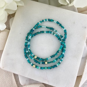 May include: Three beaded bracelets in shades of teal, turquoise, and white, with small gold beads. The bracelets are arranged in a circular pattern on a white marble surface. The beads are small and uniform in size.