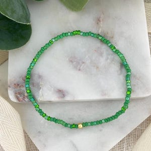May include: A beaded bracelet featuring various shades of green beads with a small gold-colored accent bead. The bracelet is circular and appears to be made of small, round beads strung together. The bracelet is displayed on a white marble surface.