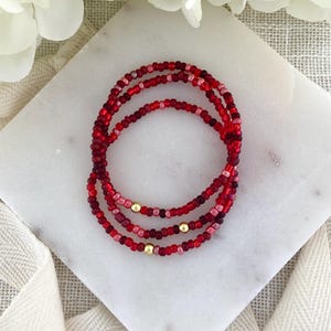 May include: Three beaded bracelets in shades of red and pink, with gold-colored accents. The bracelets are arranged in a circular pattern on a white marble surface, with a soft, neutral background.