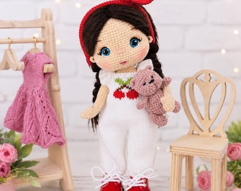 May include: A handmade crochet doll with dark brown hair in braids, wearing a red headband, a white jumpsuit with a cherry graphic, and red shoes. The doll holds a small brown crochet teddy bear. A wooden clothing rack with a pink dress stands nearby.