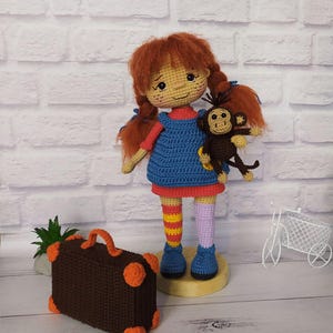 May include: A crocheted doll with red hair, a blue dress, and orange and yellow striped socks. The doll is holding a crocheted monkey. The doll is standing on a wooden base. A brown crocheted suitcase with orange accents is in the foreground.