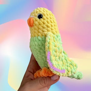 Op de afbeelding: Een handgemaakte gehaakte papegaai in geel en groen, met een oranje snavel en poten. De tekst "PENNY THE PARROT" en "design by @Purpletigerloops" zijn zichtbaar. De vogel wordt in een hand gehouden tegen een kleurrijke achtergrond.