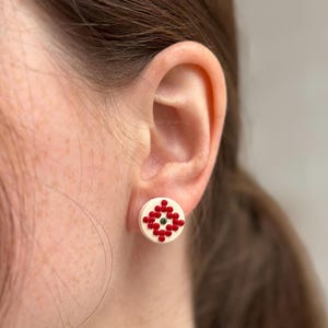 May include: A close-up of a person's ear wearing a white stud earring. The earring features a red and green geometric design. The earring is round and has a cream-colored background.