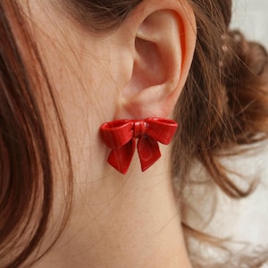 May include: A pair of red bow earrings. The earrings are made of a shiny, smooth material and are shaped like a bow. The earrings are worn by a person with brown hair.
