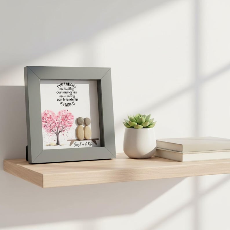 May include: A framed print with a gray border, featuring a heart-shaped pink tree and two pebble figures on a bench. The text reads "Our laughs are limitless, our memories are endless, our friendship is endless."