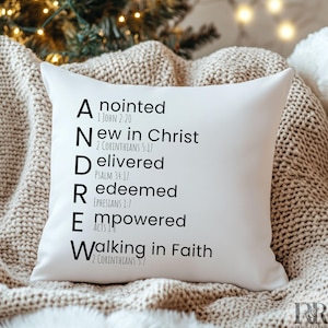 May include: A white square cushion featuring the name "ANDRE" in black, with associated words and Bible references. The cushion rests on a beige knitted blanket, suitable for home decor.