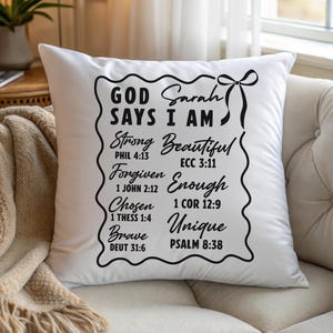 May include: White square pillow with black text that reads "GOD SAYS I AM" followed by words like "Strong, Beautiful, Forgiven, Enough, Chosen, Unique, Brave." The pillow has a decorative black border and a bow.