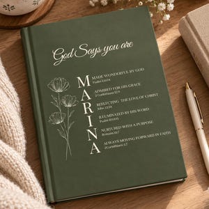Puede incluir: Un diario de tapa dura verde oscuro con el texto en blanco God Says you are y el nombre MARINA alineado verticalmente con versículos bíblicos. Un dibujo lineal de tres flores está impreso a la izquierda del nombre.