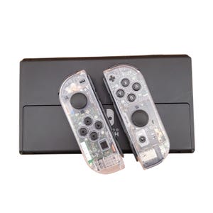 May include: Two clear, transparent Nintendo Switch Joy-Con controllers with black buttons and a black Nintendo Switch console.