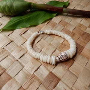 May include: A white shell bracelet with a silver clasp. The bracelet features a single, larger shell with brown spots. The bracelet is displayed on a woven, light brown surface, with green leaves and a stem in the background.