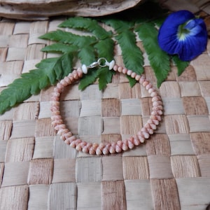 Peut inclure: Un bracelet délicat composé de petits coquillages roses clairs. Le bracelet est assemblé avec un fermoir argenté et repose sur un tapis tissé avec une fougère verte et une fleur bleue.