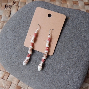 May include: A pair of dangle earrings with a natural, beachy aesthetic. Each earring features a series of white, patterned shell beads interspersed with small, pink beads. The earrings hang from silver-colored hooks and are displayed on a brown tag.