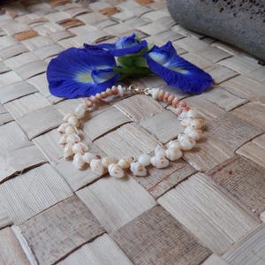 Peut inclure: Un bracelet délicat composé de petits coquillages crème, enfilés avec un fermoir doré. Le bracelet est présenté sur un tapis tissé, avec des fleurs bleues vibrantes et une pierre en arrière-plan. Le bracelet est un bijou.