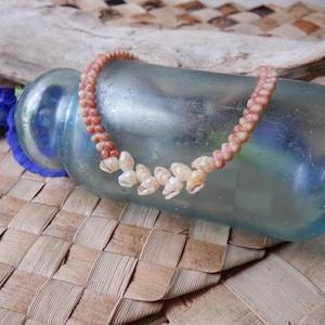 Peut inclure: Un collier de coquillages fait à la main avec un design tressé. Le collier présente de petits coquillages de couleur claire et un cordon tressé dans des tons pêche et crème. Le collier est présenté sur une bouteille en verre bleu vintage.