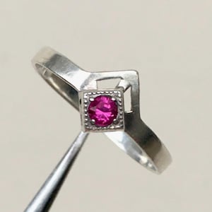 May include: A silver ring with a square setting featuring a vibrant pink gemstone. The ring has a unique, angular design with a polished finish. The gemstone is the focal point, set within a detailed square frame.