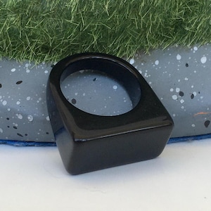 Könnte beinhalten: Ein glänzender schwarzer Ring mit rechteckiger Oberfläche und abgerundeter Innenseite. Der Ring besteht aus einem soliden, dunklen Material und liegt auf einer weißen Oberfläche. Das Design des Rings ist schlicht und modern.