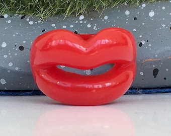 May include: A glossy, red lip-shaped ring. The ring is a bold, statement piece, with a smooth, reflective surface. The design is a playful and eye-catching accessory, perfect for adding a pop of color and personality to any outfit.