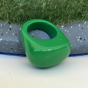 May include: A vibrant green ring with a smooth, glossy finish. The ring has a rounded, geometric shape, and is set against a neutral background. This bold accessory is a statement piece.
