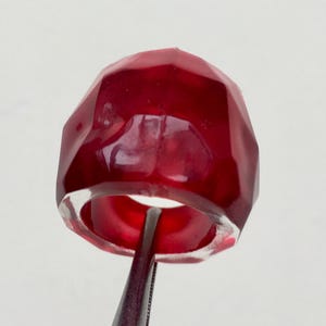 May include: A deep red, faceted ring made of resin. The ring is thick and has a wide band.