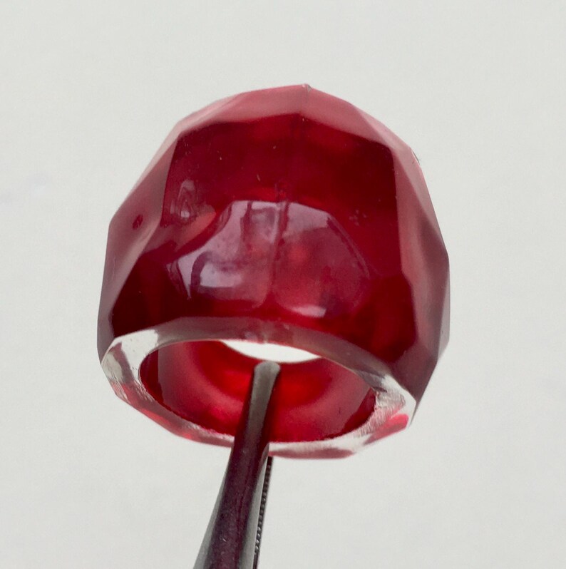 May include: A deep red, faceted ring made of resin. The ring is thick and has a wide band.