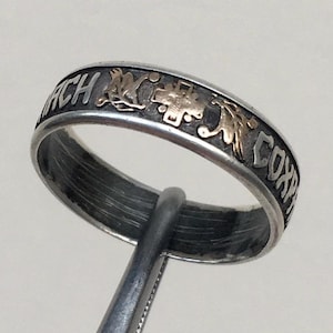 May include: Silver ring with gold accents and engraved text. The ring features a band with alternating text and decorative gold elements. The ring is held by a metal tool.