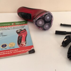 May include: A red electric shaver with a rotating head and a black power cord. The shaver is on a white surface with a manual in Ukrainian.