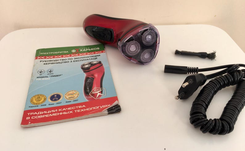 May include: A red electric shaver with a rotating head and a black power cord. The shaver is on a white surface with a manual in Ukrainian.