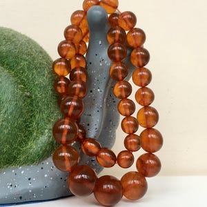 Puede incluir: Una larga hebra de cuentas de &aacute;mbar, con un color c&aacute;lido y mielado. Las cuentas son redondas y lisas, y est&aacute;n enhebradas en un cord&oacute;n delgado. Las cuentas est&aacute;n dispuestas en una doble hebra, y est&aacute;n drapeadas sobre una superficie gris texturizada.