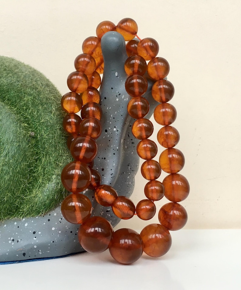 Puede incluir: Una larga hebra de cuentas de &aacute;mbar, con un color c&aacute;lido y mielado. Las cuentas son redondas y lisas, y est&aacute;n enhebradas en un cord&oacute;n delgado. Las cuentas est&aacute;n dispuestas en una doble hebra, y est&aacute;n drapeadas sobre una superficie gris texturizada.