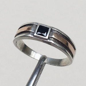 Op de afbeelding: Een zilveren ring met een vierkante zwarte edelsteen in het midden. De ring heeft een goudkleurige band en een getextureerd ontwerp aan de zijkanten. De ring wordt vastgehouden door een metalen gereedschap.
