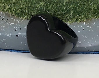 Può includere: Un anello nero lucido a forma di cuore. L'anello è realizzato in un materiale solido e opaco con una superficie liscia e lucida. È posto su uno sfondo di superficie grigia macchiata e erba verde.