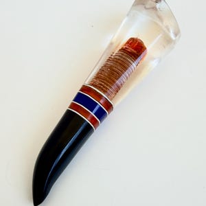 May include: A tapered handle featuring a clear resin top section that encases a textured, cylindrical copper-colored core. The lower portion is solid black, separated from the top by a decorative band of alternating blue and copper stripes.