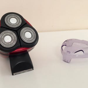 May include: A red electric razor with three rotating blades. The razor has a black handle and a clear plastic cover.