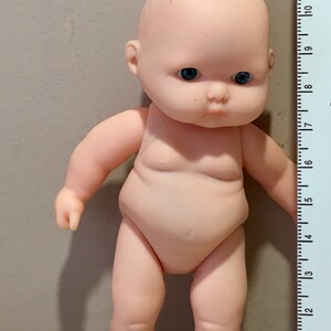 May include: A small, nude-colored plastic baby doll with blue eyes. The doll is about 6 inches tall and is standing next to a ruler.