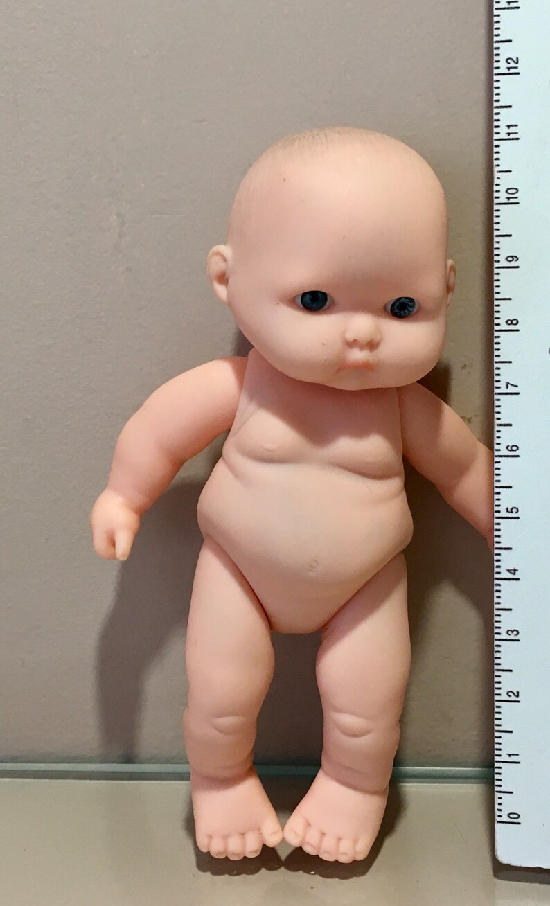 May include: A small, nude-colored plastic baby doll with blue eyes. The doll is about 6 inches tall and is standing next to a ruler.