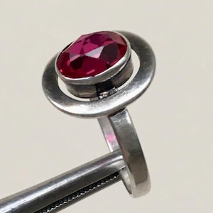 May include: A silver ring with a deep red faceted gemstone set in a double circle design.