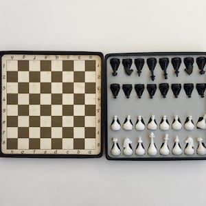 Op de afbeelding: Een vintage schaakset met een zwart-wit geblokt bord en zwarte en witte schaakstukken. De set zit in een zwarte koffer, met het bord aan de ene kant en de stukken in een bakje aan de andere kant.