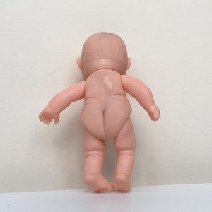 May include: A small, nude-colored plastic baby doll with a painted face and no clothing. The doll is standing with its back to the camera.