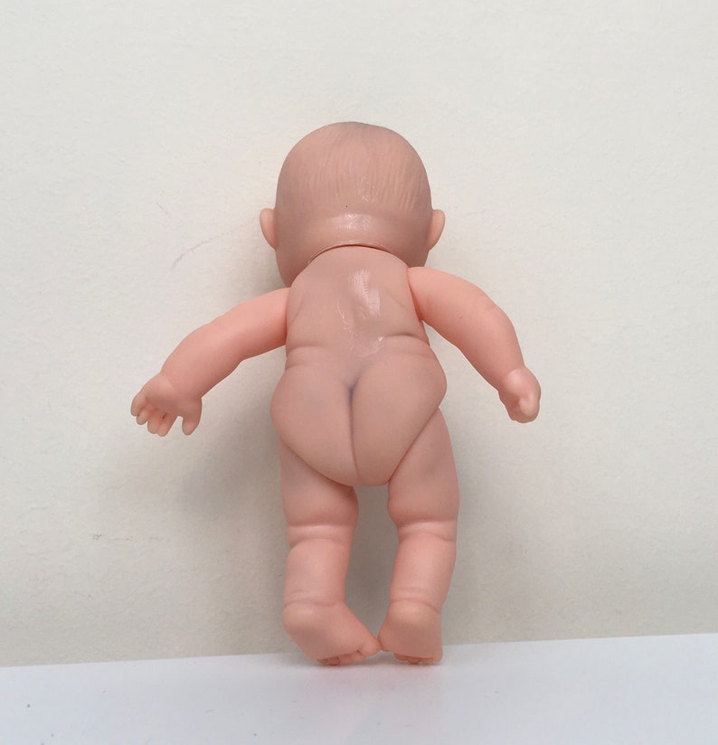 May include: A small, nude-colored plastic baby doll with a painted face and no clothing. The doll is standing with its back to the camera.