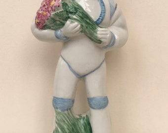May include: A white ceramic figurine of an astronaut holding a bouquet of pink and green flowers. The astronaut is wearing a white spacesuit with blue accents and a white helmet with a gold trim.