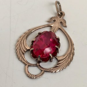 Könnte beinhalten: Ein Vintage-Anhänger mit einem großen, ovalen, rubinroten Edelstein, der in einem dekorativen, goldfarbenen Metallrahmen gefasst ist. Der Rahmen hat ein verziertes, offenes Design mit Blatt- und Scroll-Details und eine kleine Schlaufe für eine Kette.