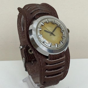 Puede incluir: Un reloj de pulsera plateado con una esfera blanca y agujas negras. El reloj está en una correa de cuero marrón.