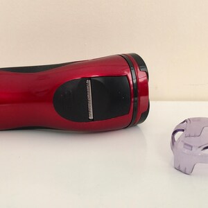May include: A red electric razor with a black handle and a clear plastic guard. The razor has a silver trim and the text "HK-6885 220V-240V 50Hz 18 watts" printed on the side.