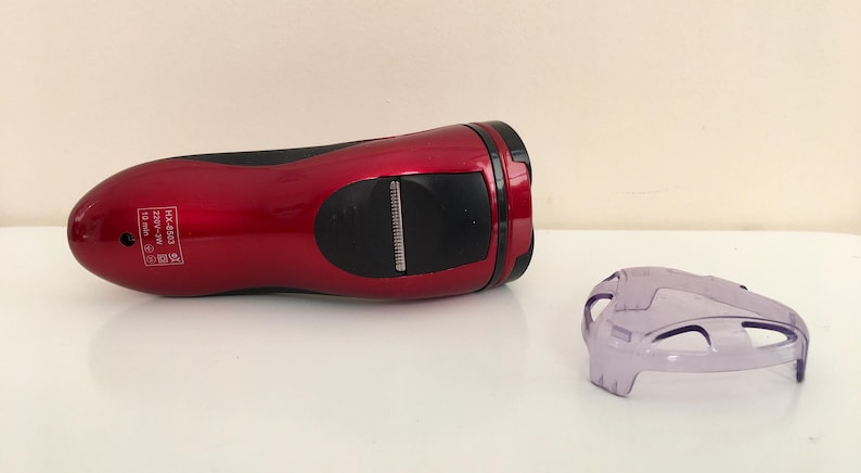 May include: A red electric razor with a black handle and a clear plastic guard. The razor has a silver trim and the text "HK-6885 220V-240V 50Hz 18 watts" printed on the side.