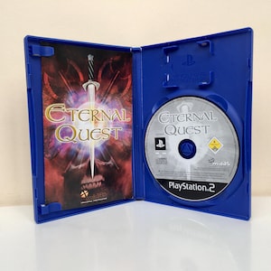 Puede incluir: Una caja de juego de PlayStation 2 azul con un disco plateado en el interior. El juego se llama "Eternal Quest" y presenta una espada y una calavera en la portada.
