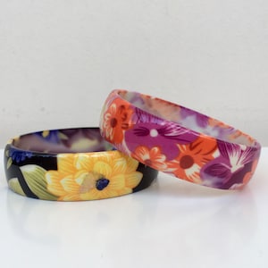 Puede incluir: Dos pulseras de plástico con estampado floral. La pulsera superior es de color morado, naranja y blanco con un estampado floral. La pulsera inferior es de color negro, amarillo, verde y morado con un estampado floral.