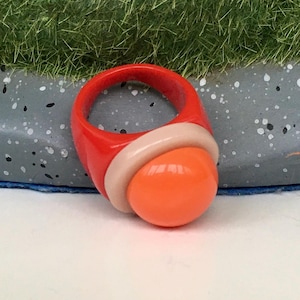 May include: A striking ring with a retro aesthetic. It features a bright red band, a light beige accent, and a prominent orange orb. The design is bold and modern, perfect for adding a splash of colour to any outfit. The ring is a statement piece.