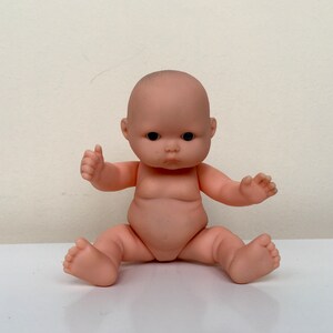 May include: A small, nude-colored baby doll with black eyes and a realistic face. The doll is sitting with its legs crossed and its arms outstretched.
